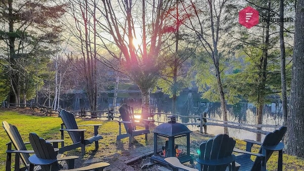 Un espacio tranquilo al aire libre con sillas Adirondack y un brasero, rodeado de árboles y vistas a un cuerpo de agua.