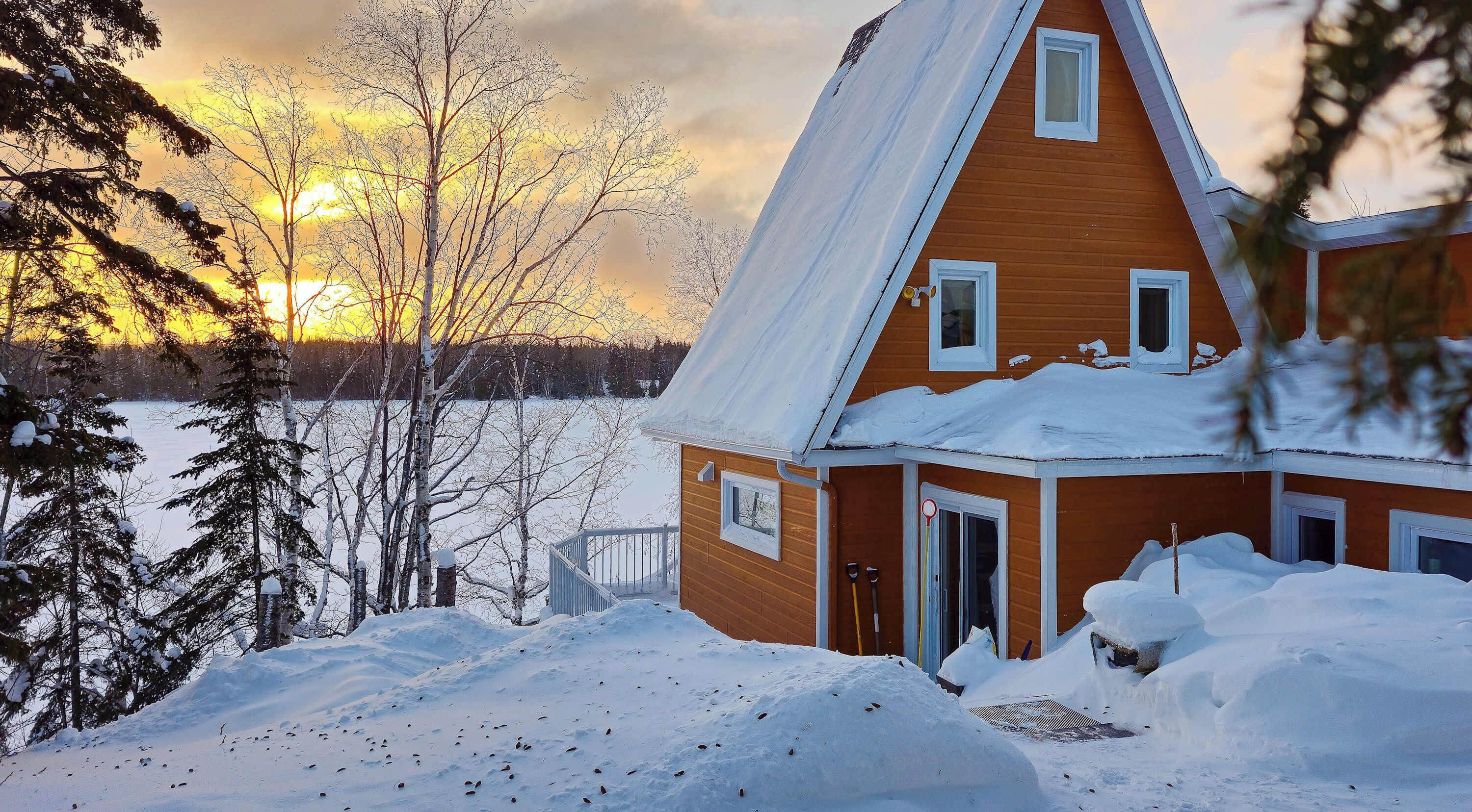 A wooden house with a steep roof, surrounded by snow-covered ground and trees, with a view of the sunset in the background.