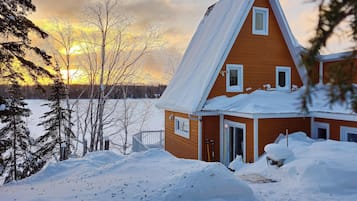 A wooden house with a steep roof, surrounded by snow-covered ground and trees, with a view of the sunset in the background.