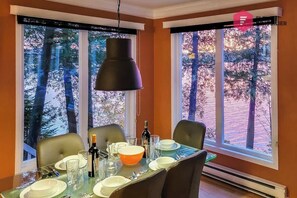 A dining area with a table set for four, large windows with a view of trees, and a black pendant light. - Aux Berges du Pont Suspendu - Grand Lac Brompton (Saint-Denis-de-Brompton)