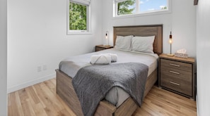 A bedroom with a wooden bed, two bedside tables, a nightstand, a dresser, and a window with a view of trees.