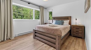 A bedroom with a wooden bed, a nightstand, a window with curtains, and a view of trees.