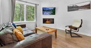 A living room with a leather sofa, a wooden coffee table, a recliner, a fireplace, and a large window with a view of trees.