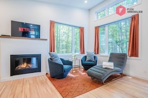 A modern living room with a fireplace, two armchairs, a sofa, a TV, and large windows with curtains.