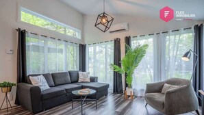 A modern living room with a black sofa, a round coffee table, and a grey armchair. There is a large window with curtains, a potted plant, and a hanging light fixture. - The Palm Tree of Portneuf - Private SPA Pool Trail (Pont-Rouge)