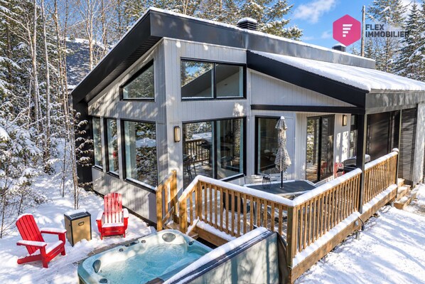 Una casa moderna de dos pisos con un balcón, un jacuzzi y un paisaje cubierto de nieve.