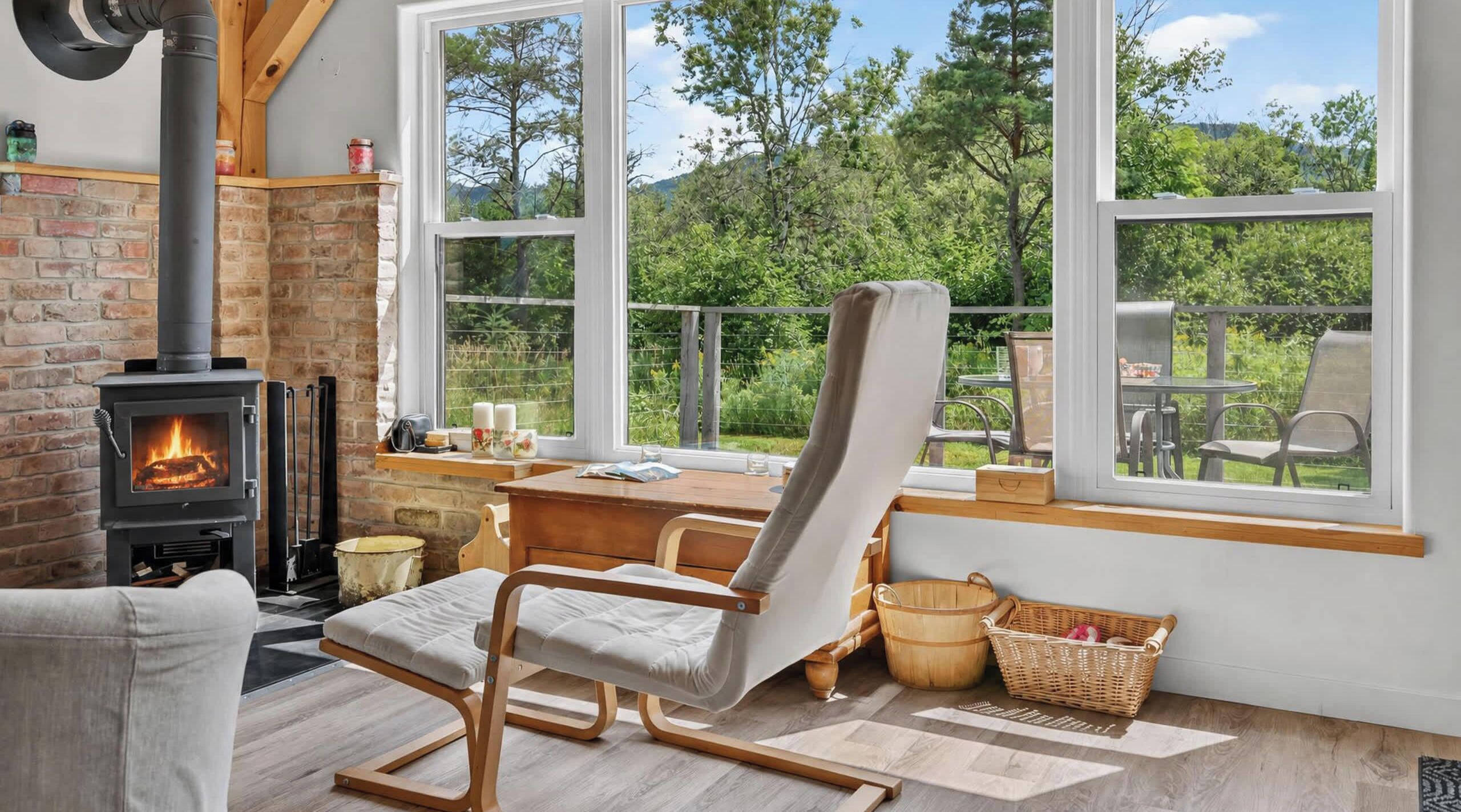 Un salon chaleureux avec une cheminée, un fauteuil confortable, une table à manger et de grandes fenêtres donnant sur l’extérieur.