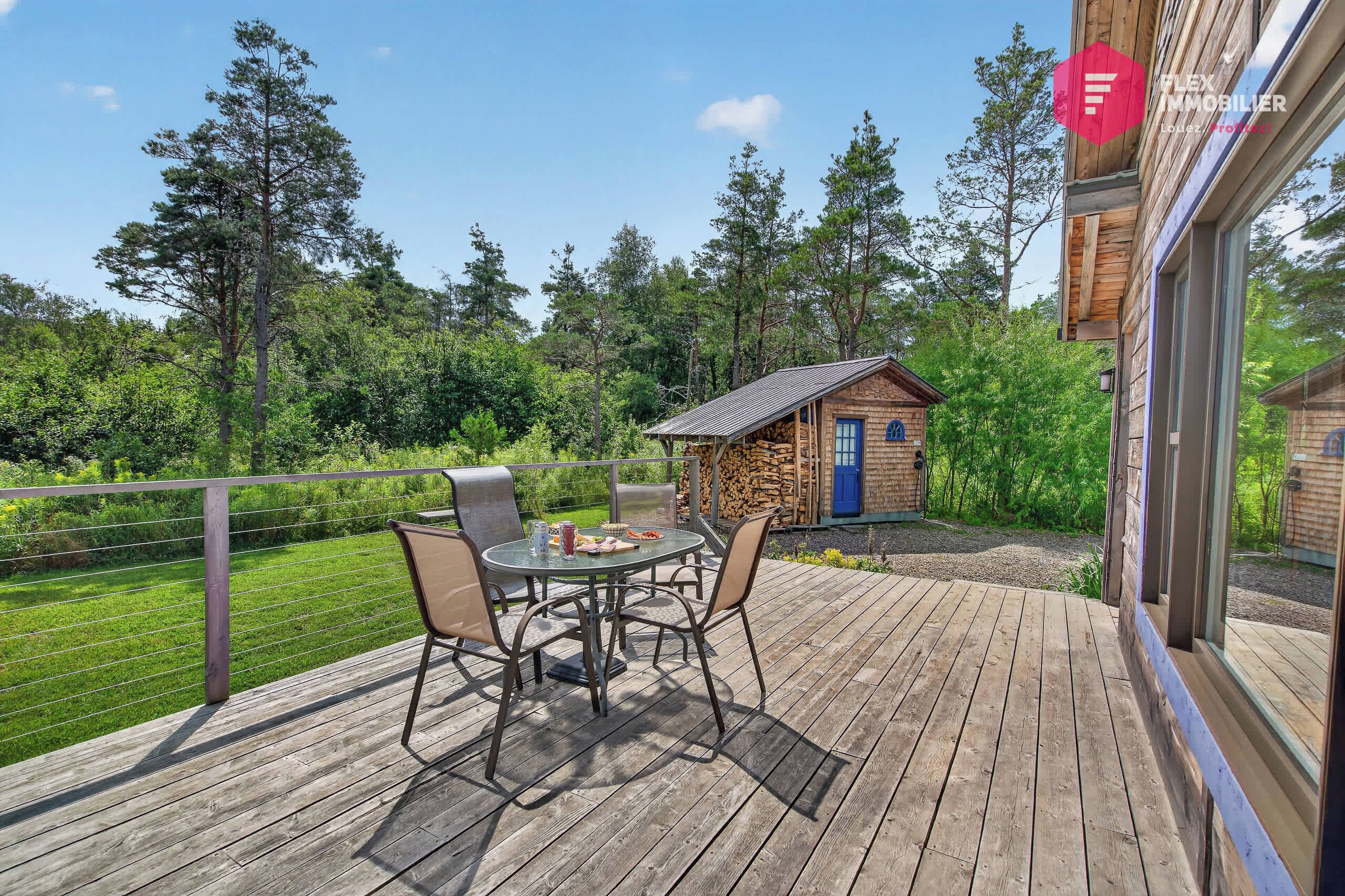 A wooden deck with a table and chairs, a small building with a blue door, and a view of a forested area.