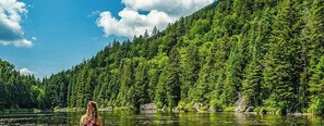 Une femme est assise au bord d’une rivière, avec une montagne boisée en arrière-plan.