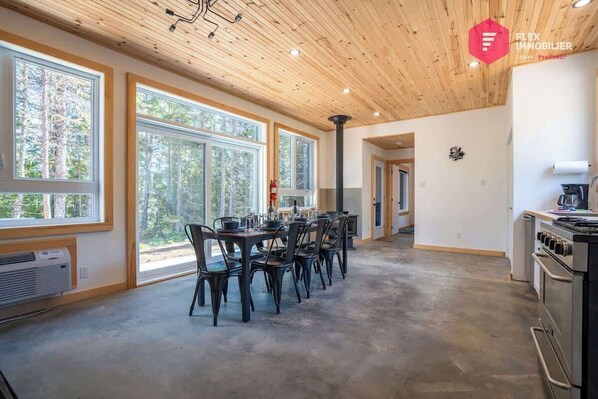 Una habitación amplia con ventanas grandes, un comedor con sillas, una cocina con estufa y un techo con vigas de madera.