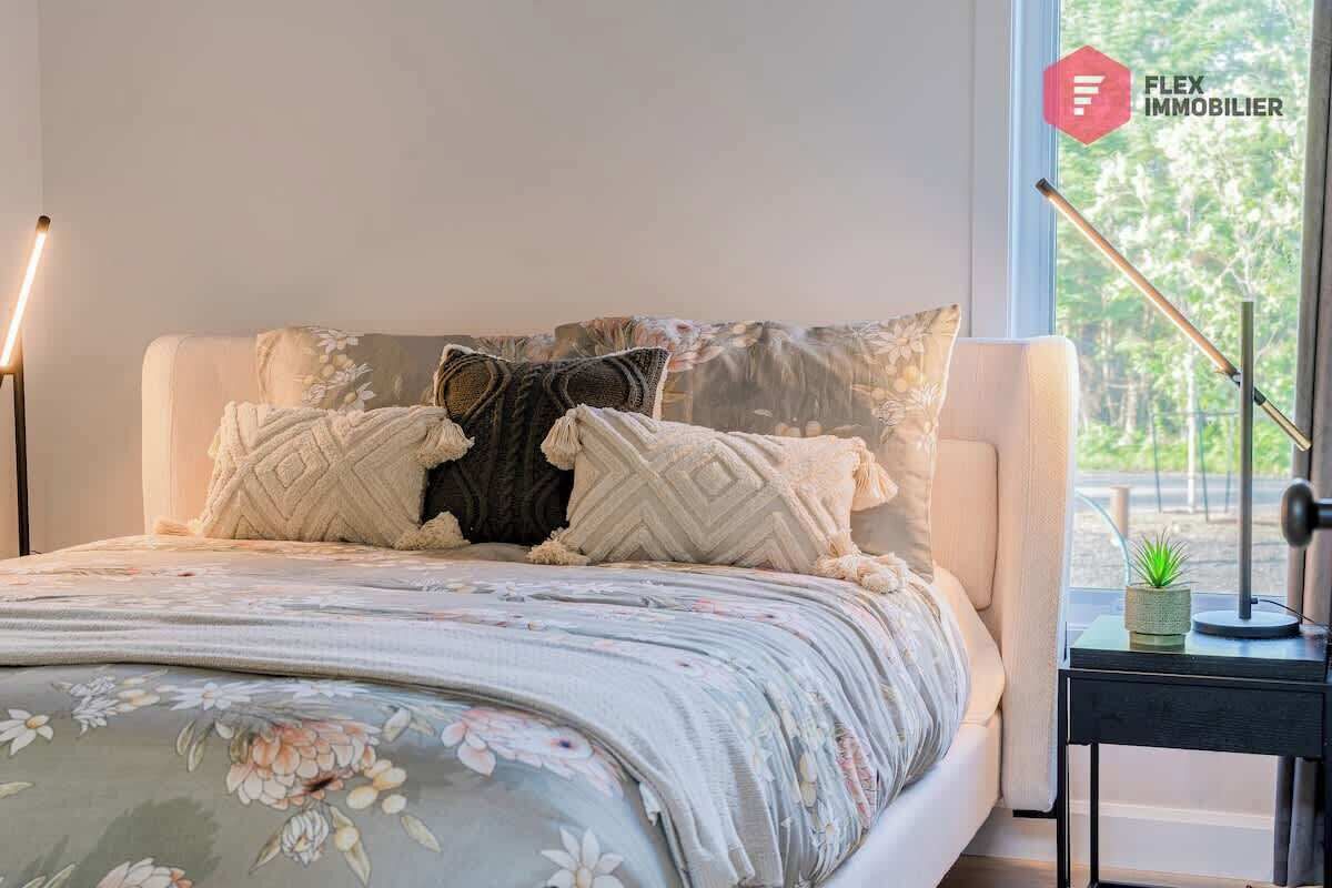 Een slaapkamer met een bed, kussens, een nachtkastje, een lamp en een raam met uitzicht op bomen.