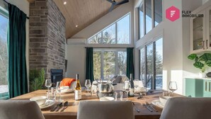 Un coin repas avec une table en bois dressée pour quatre personnes, une cheminée en pierre et de grandes fenêtres donnant sur la forêt.