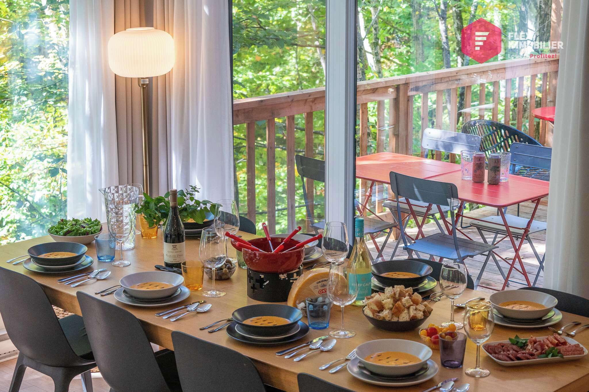 Un espacio de comedor al aire libre con una mesa de madera lista para una comida, rodeado de sillas y vistas a un área boscosa.