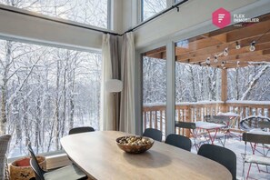 A dining area with a large table and chairs, a bowl of fruit, and a view of a snowy forest through large windows.