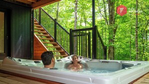 A hot tub with two people, a wooden staircase, and a forested background. - The Bellevue in Portneuf - Private SPA, 65-inch TV (Pont-Rouge)