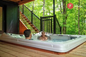 Ein Whirlpool mit zwei Personen, einer Holztreppe und einem bewaldeten Hintergrund.