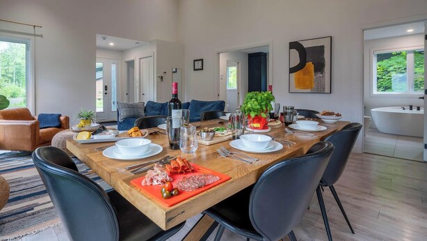 A dining area with a wooden table set for four, surrounded by chairs, with a view of a modern bathroom and a large window. - The Bellevue in Portneuf - Private SPA, 65-inch TV (Pont-Rouge)
