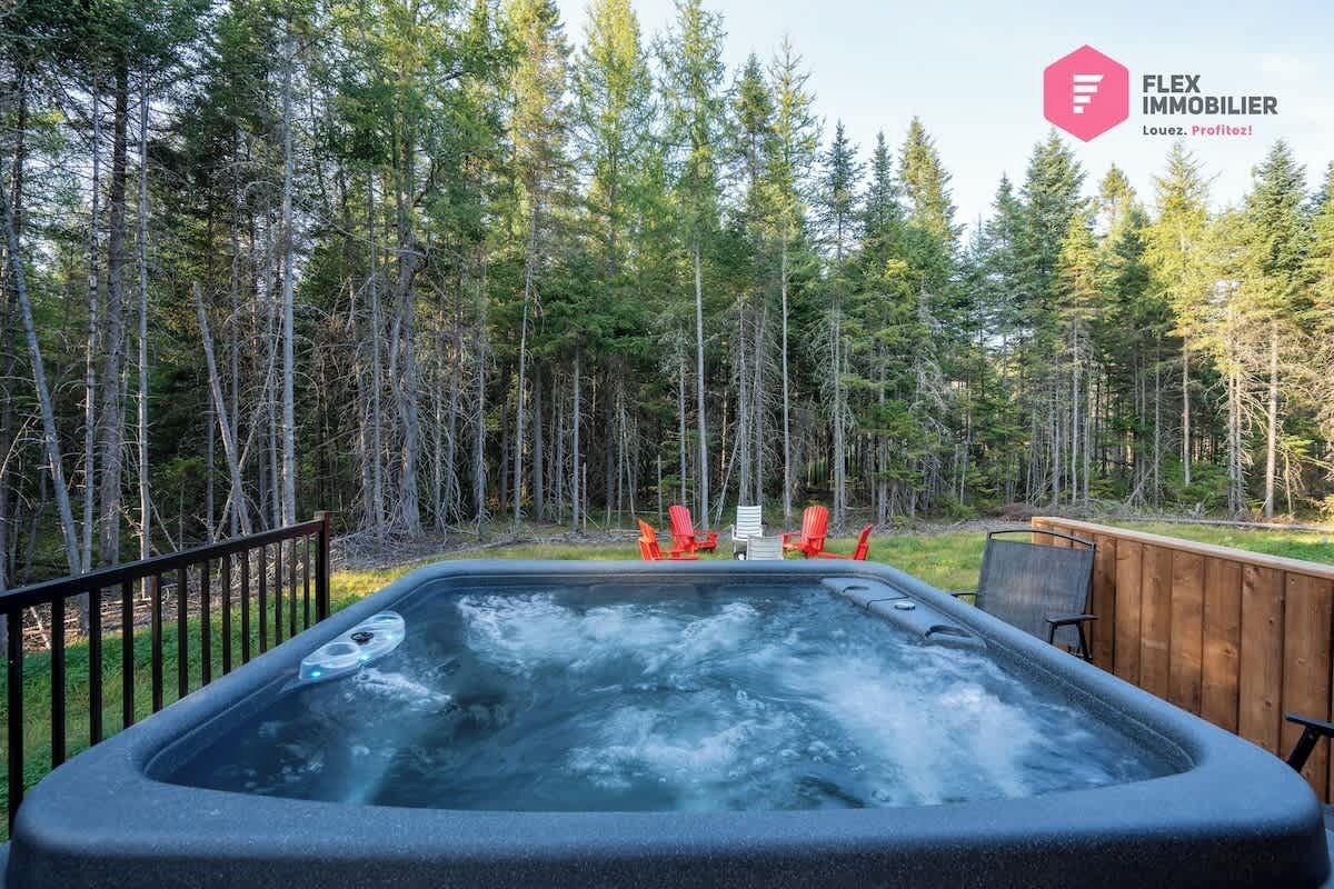 Een hottub omgeven door een houten terras met rode stoelen, met een bos op de achtergrond.