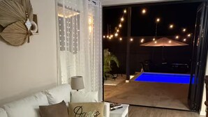 Ein Wohnzimmer mit einem weißen Sofa, einem Couchtisch und einem Kissen mit dem Schriftzug "Oasis". Eine Glastür führt zu einem Außenbereich mit Lichterketten und einem Pool.