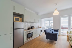 A modern kitchen with white cabinets, a microwave, a refrigerator, and a washing machine. There is a blue chair and a table with a floral pattern.
