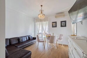 A modern living room with a brown leather sofa, a glass dining table with chairs, a chandelier, and a flat-screen TV mounted on the wall. - Lovely Urbisado III -  By Y Concept (Setúbal)