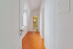 A narrow hallway with white walls and orange-tiled flooring leading to a room with a circular window. - Sapalinho Retreat B - By Y Concept (Setúbal)