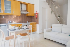 A modern kitchen with wooden cabinets, a dining area with white chairs, and a staircase leading to a living area with a white sofa. - Setubal Downtown - By Y Concept (Setúbal)