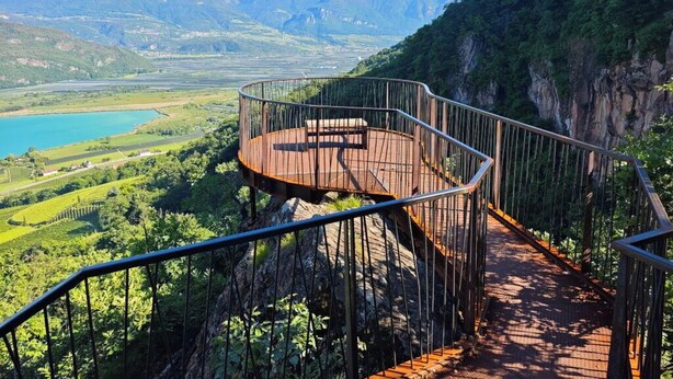 Una escalera de caracol con barandilla de metal que lleva a una plataforma de madera con vistas a un valle, un lago y montañas.