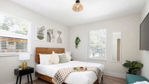A bedroom with a wooden bed, a bedside table with a lamp, a wall-mounted TV, and a window with blinds. - Serene Place, Walk to UF, Stadium, Near Shands & Downtown (Gainesville)