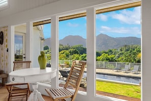 A room with a white table and chairs, a large window with a view of mountains and greenery. - Nānā Lani, Artsy Escape with Inspiring Views! (Princeville)