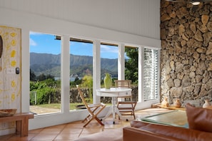 A room with large windows offering a view of mountains and greenery. There is a round table with chairs, a bench, and a stone wall. - Nānā Lani, Artsy Escape with Inspiring Views! (Princeville)
