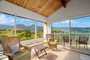 A balcony with wicker chairs, a wooden table, and a view of mountains and greenery. - Nānā Lani, Artsy Escape with Inspiring Views! (Princeville)