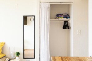 A room with a wooden table, a mirror, a small potted plant, a yellow chair, a white curtain, and a shelf with folded clothes and a toy train. - Cozy Island Stay-Waikiki (Honolulu)