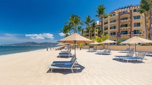 Ein Strand mit Liegestühlen und Sonnenschirmen, ein Gebäude mit Balkonen und Palmen.