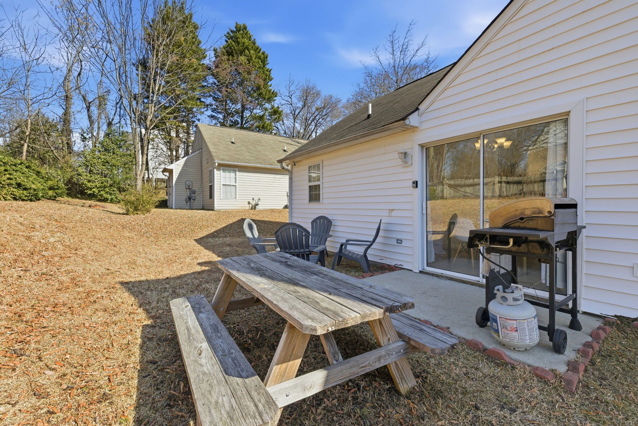 A wooden picnic table with chairs, a grill, and a propane tank outside a house with a sliding glass door.