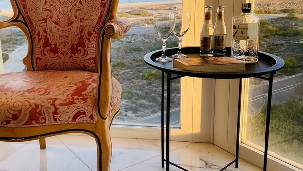 Una habitación con vistas a la playa, un sillón rojo con estampado, una mesita con vasos y botellas, y una botella de ginebra japonesa.