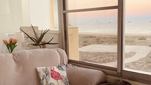 Una sala moderna con un sofá color beige, un cojín decorativo, una cesta tejida y vista a la playa a través de amplias ventanas.
