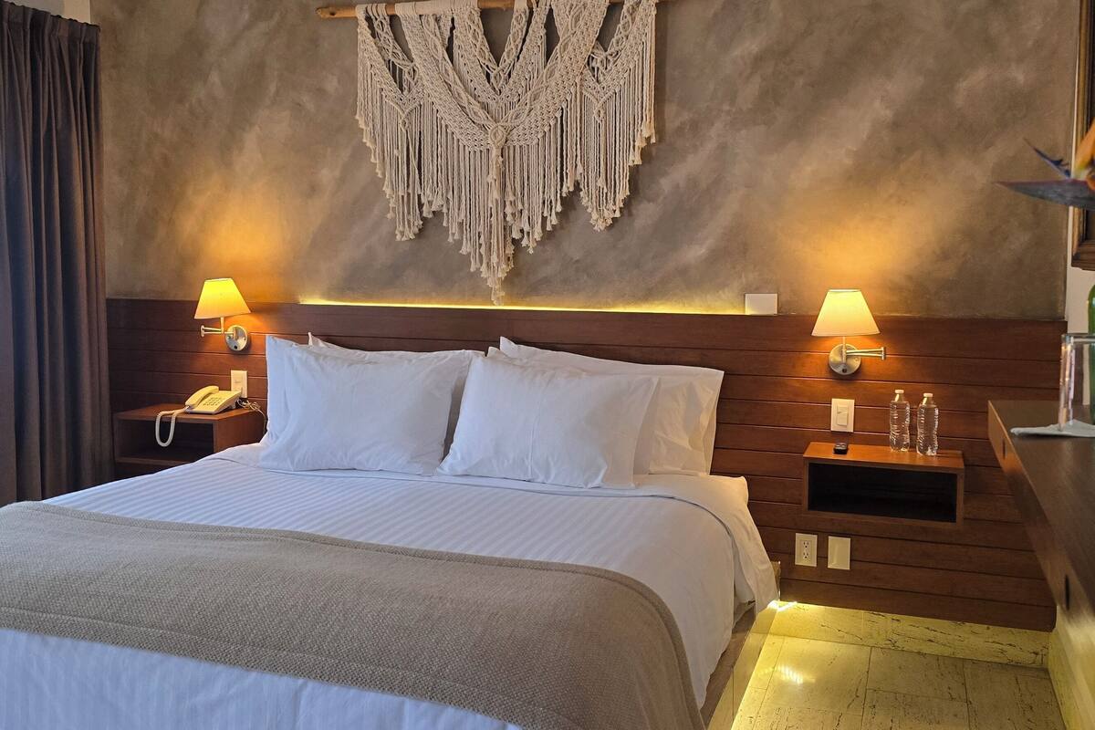 A hotel room with a large bed, two bedside tables with lamps, a macrame wall hanging, and a vase with flowers.