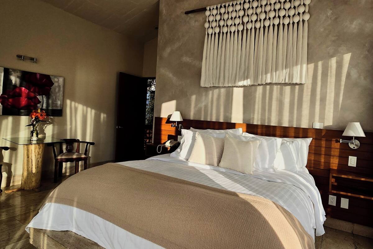 A hotel room with a large bed, a wooden headboard, a bedside table with a lamp, a small table with a vase of flowers, and a painting on the wall.