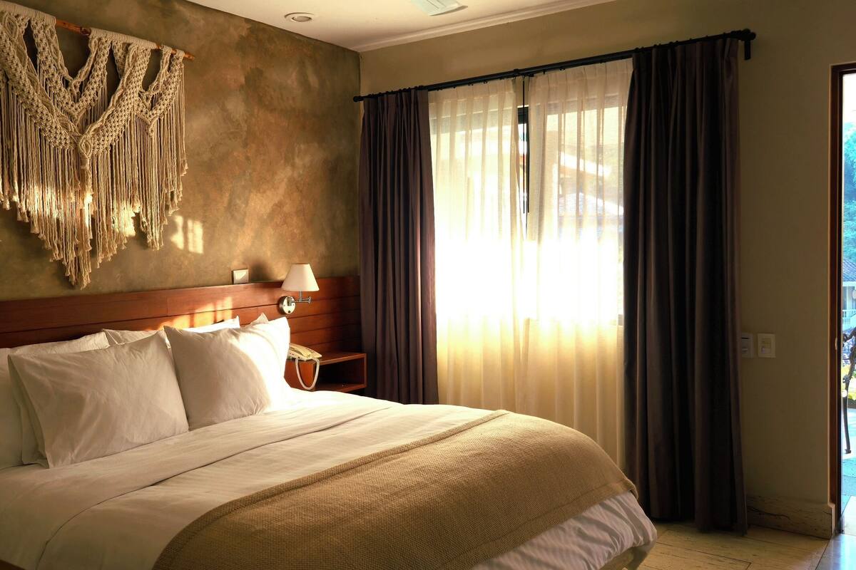 A hotel room with a large bed, a macrame wall hanging, a bedside table with a lamp, and a view of a garden through sheer curtains.