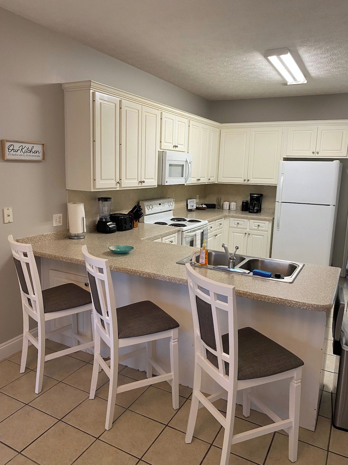 Una cocina con gabinetes blancos, microondas, estufa, refrigerador y dos taburetes de bar con asientos acolchados.