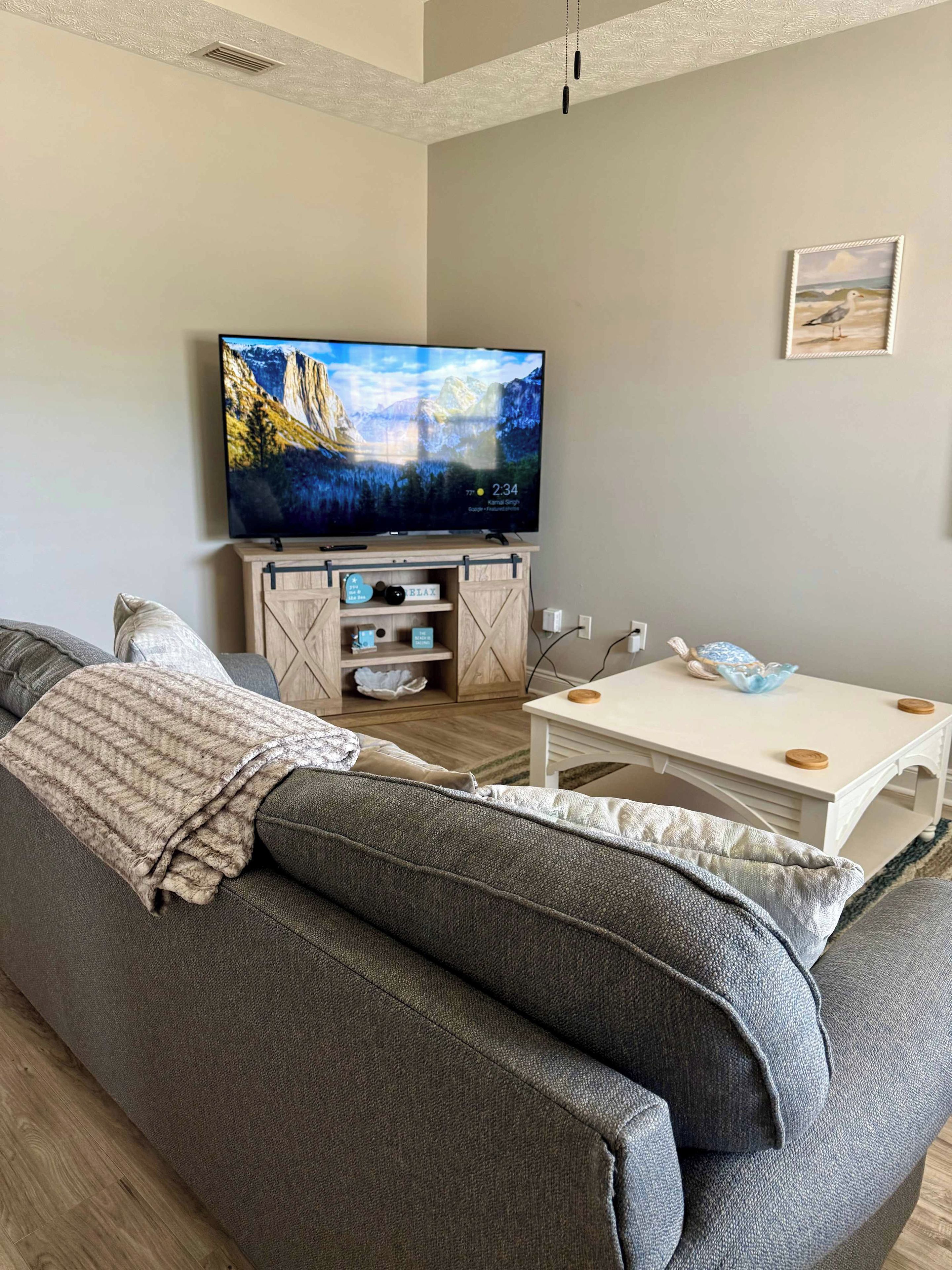 Een woonkamer met een groot flatscreen televisietoestel op een houten standaard, een grijze bank, een witte salontafel en een kleine witte tafel met een lamp.