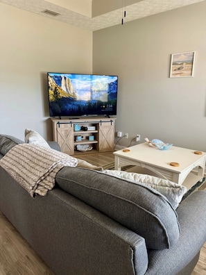 Een woonkamer met een groot flatscreen televisietoestel op een houten standaard, een grijze bank, een witte salontafel en een kleine witte tafel met een lamp.