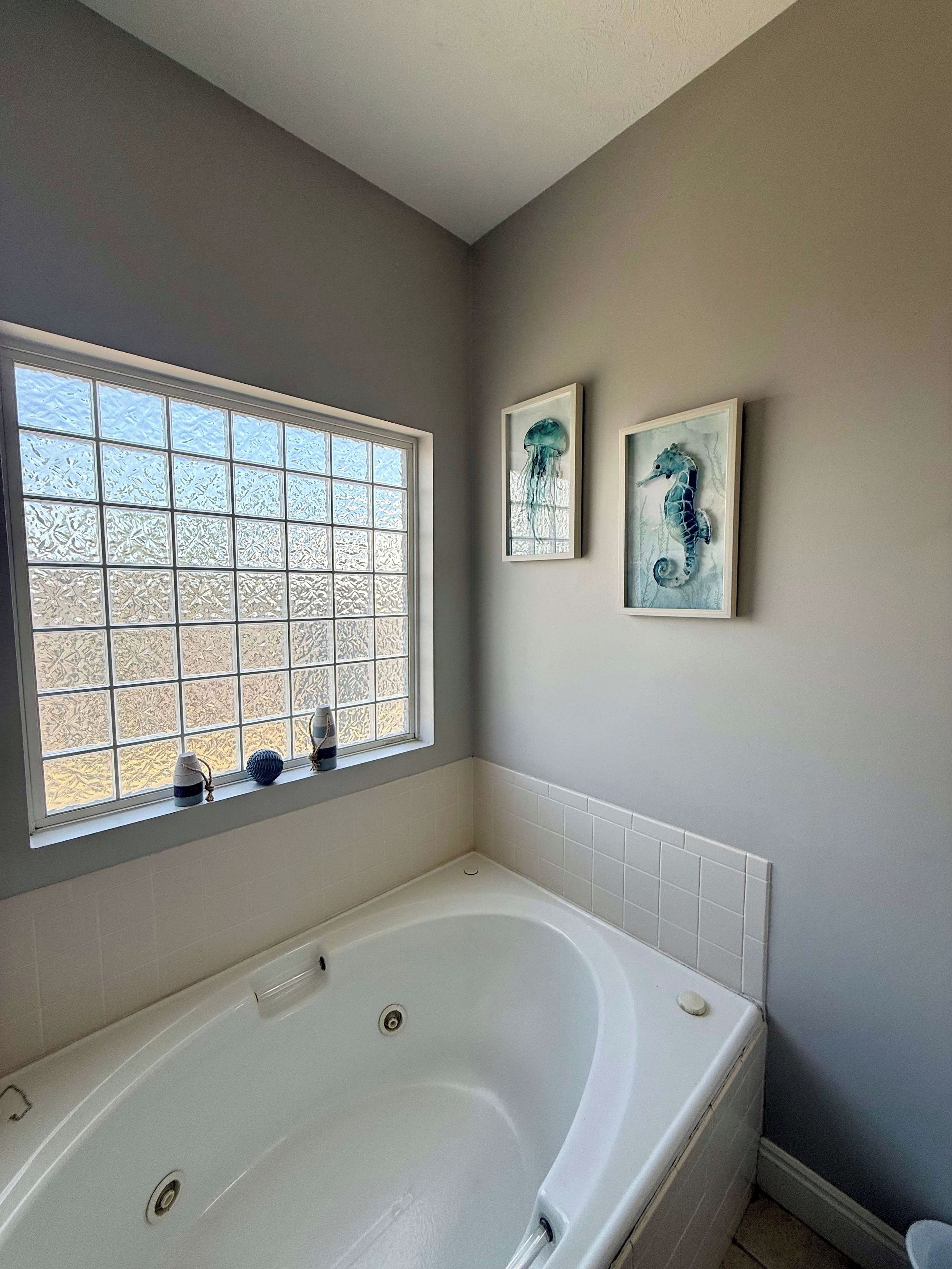 A bathroom with a large bathtub, frosted window, and two framed artworks on the wall.
