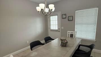 A dining area with a long table, black chairs, a chandelier, and a window with blinds.