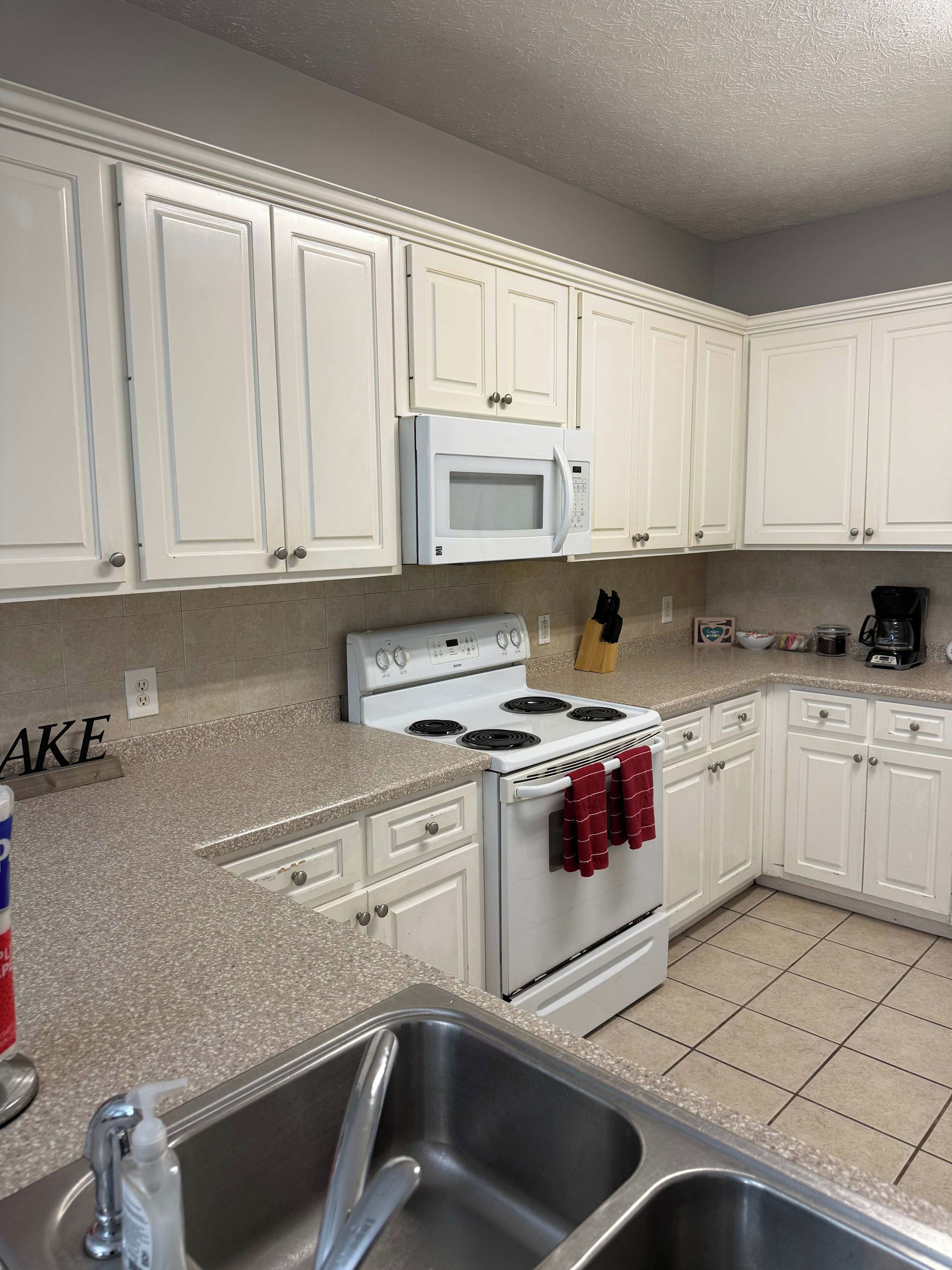 A kitchen with white cabinets, a white microwave, a stove, and a stainless steel sink.