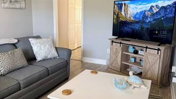A living room with a grey sofa, a white coffee table with wooden coasters, a television on a wooden stand, and a patterned rug.