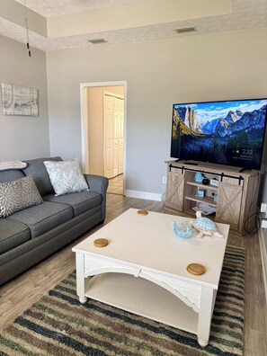 A living room with a grey sofa, a white coffee table with wooden coasters, a television on a wooden stand, and a patterned rug. - Beach Days, Nightlife Nights, East End of PCB (Panama City)