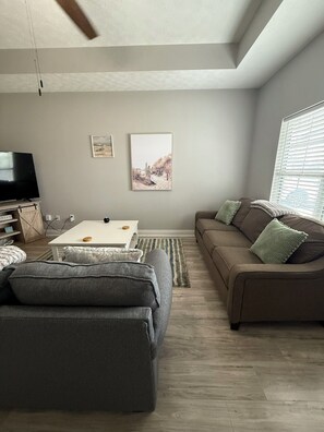 Ein modernes Wohnzimmer mit grauem Sofa, einem weißen Couchtisch und einem braunen Sofa mit grünen Kissen. Ein Flachbildfernseher steht auf einem Holzboden, an der Wand hängt ein gerahmtes Bild und es gibt ein Fenster mit Jalousien.