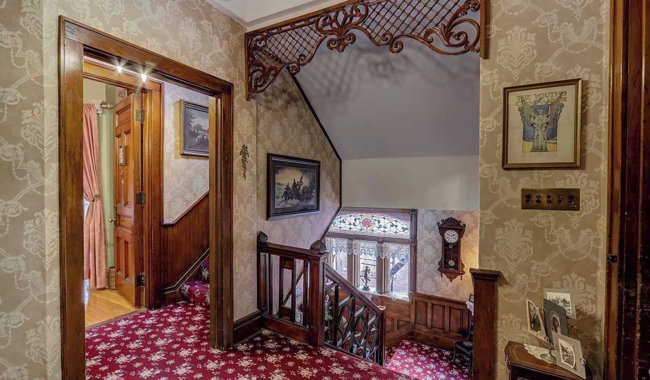 A staircase with ornate railings and patterned wallpaper, leading to a landing with a clock and framed pictures.